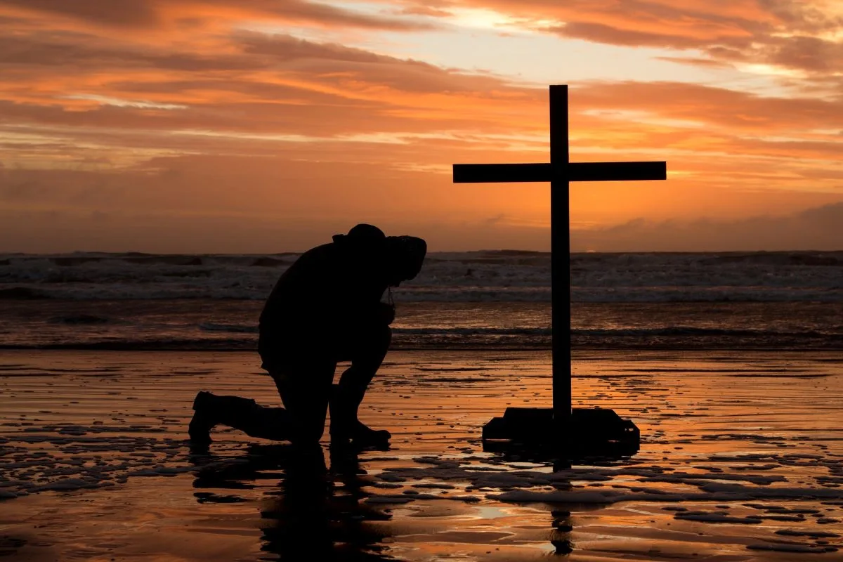 Man kneels in front of cross at sunset.