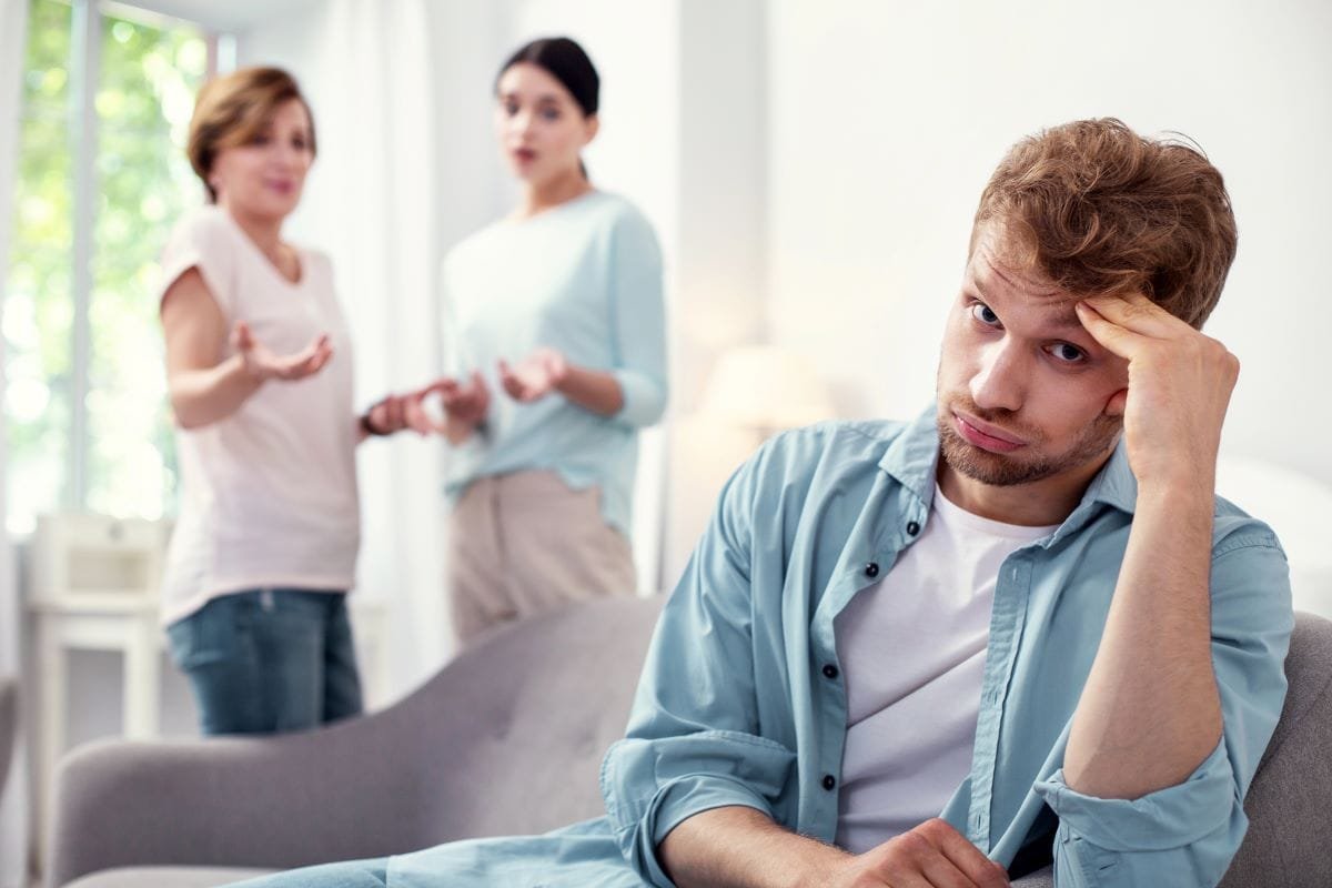 A man sits exasperated with two women behind him looking at him.
